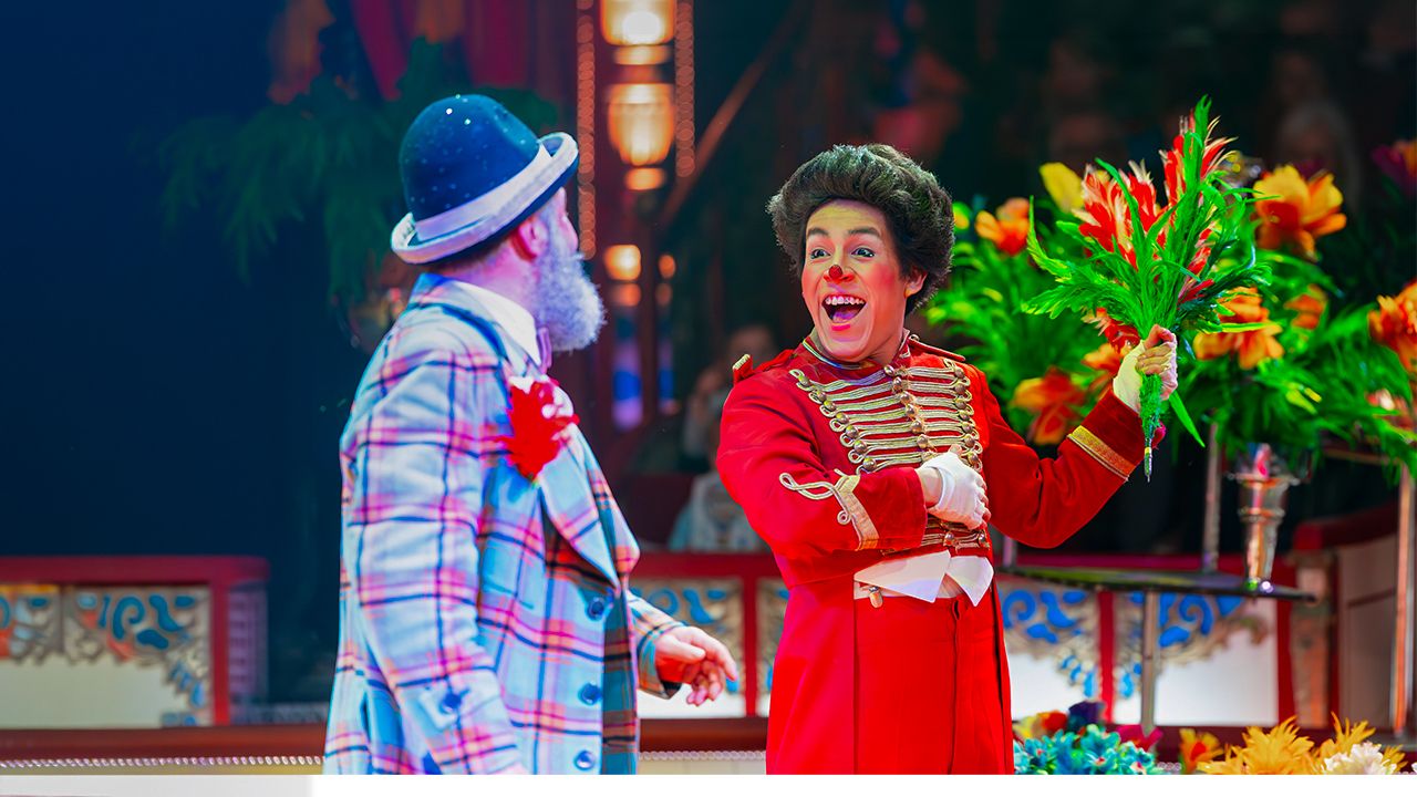 Ein Clown mit einem bunten Blumenstrauß steht einem anderem Clown in der Manege gegenüber und lacht