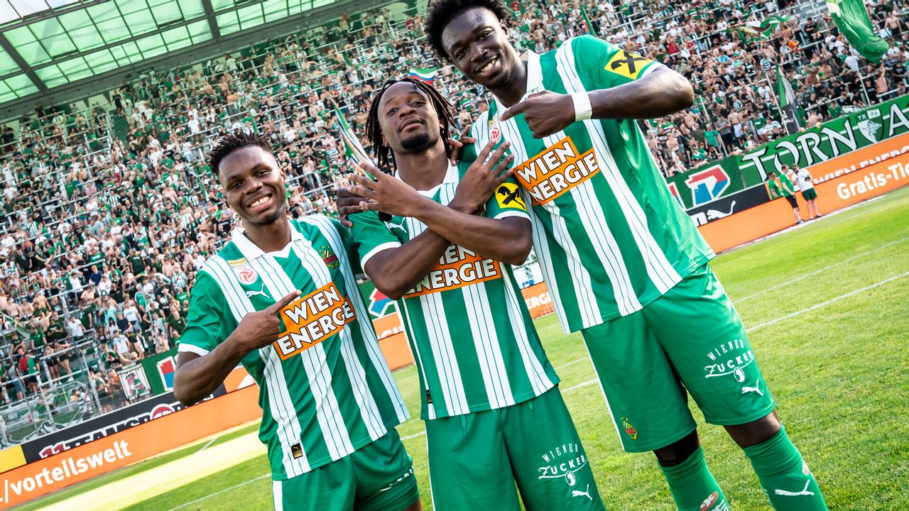 Drei Fußballspieler in Rapid-Dressen am Feld vor Publikum