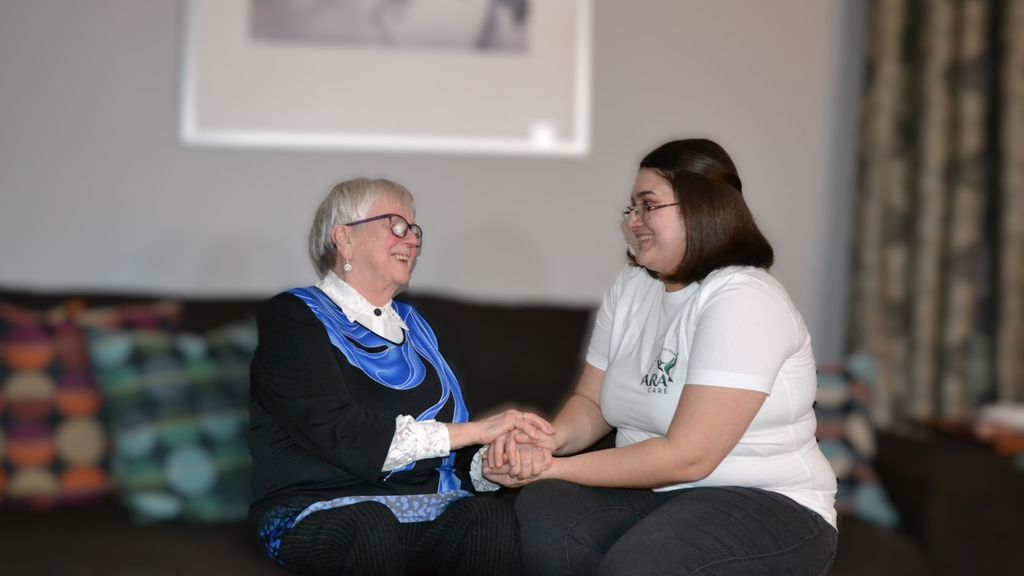 Pflegerin mit einer älteren Dame auf einer Couch sitzend