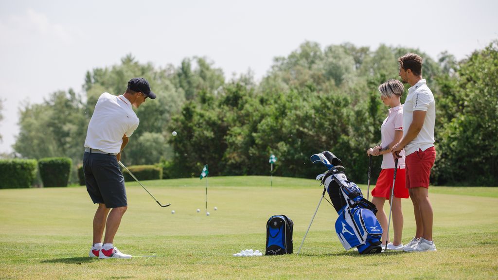 Ein Golftrainer mit einem Pärchen, welches gerade Golfen lernen möchte stehen am Golfplatz. 
