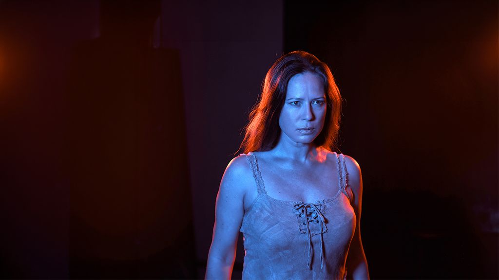 Frau mit langen Haaren steht im rot-blau beleuchteten Bühnenlicht und blickt ernst nach vorne.