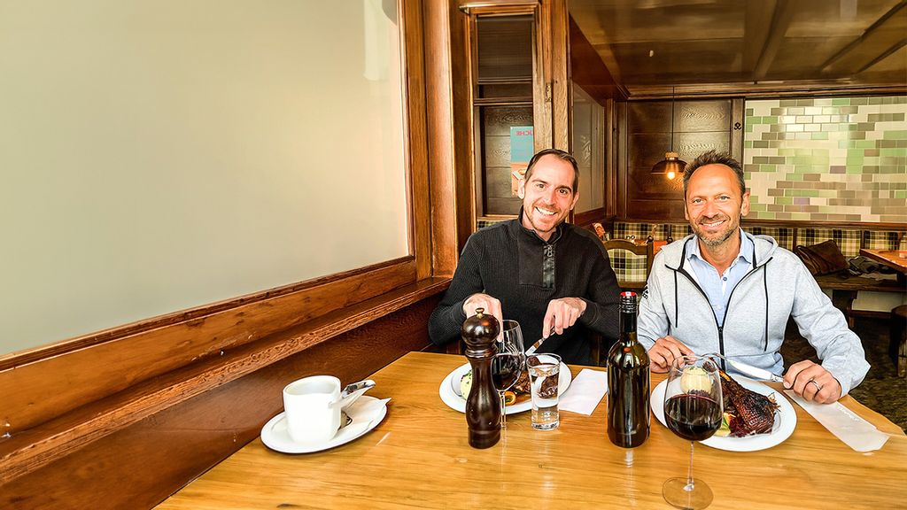 Zwei Männer sitzen mit Speisen und Getränken an einem Tisch und lächeln