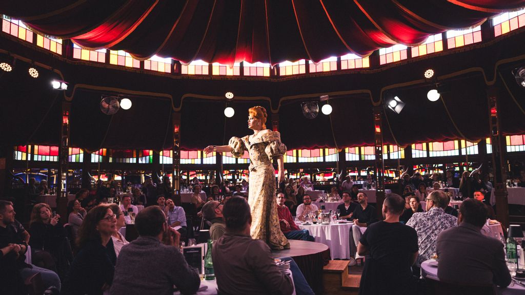 Drag Queen steht in der Mitte eines gefüllten Saales bei einer Brunch-Show 