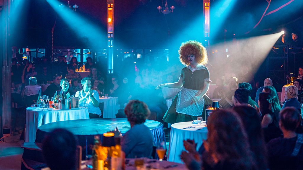 Drag Queen steht in der Mitte eines gefüllten Saales bei einer Brunch-Show 