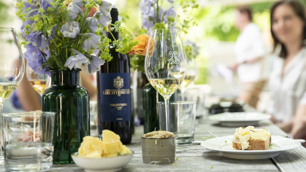 Ein leicht gefülltes Weinglas steht neben einer Weinflasche, Speisen und einer Blumenvase