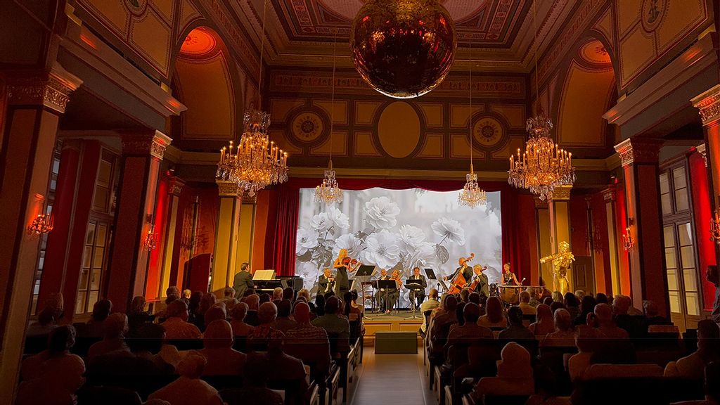 Orchester mit diversen Streichinstrumenten auf der Bühne, davor der Dirigent in schwarzem Anzug