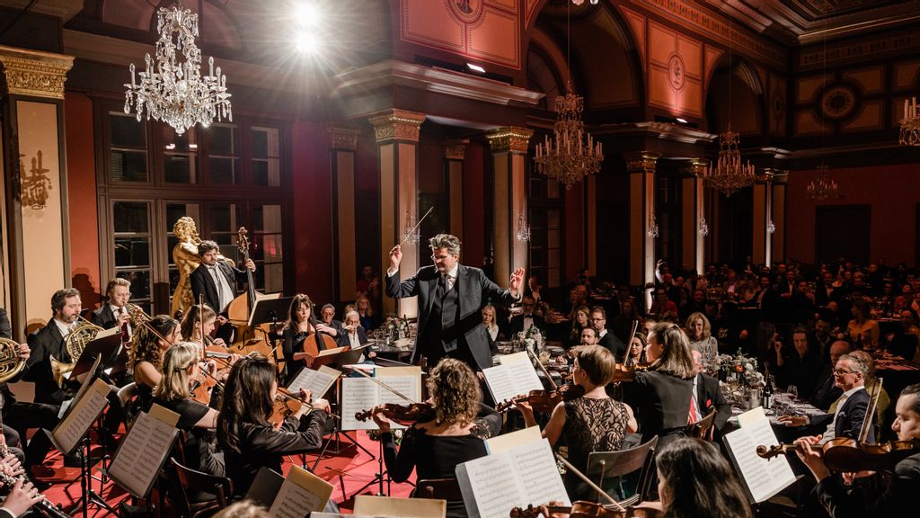 Orchester mit diversen Streichinstrumenten auf der Bühne, davor der Dirigent in schwarzem Anzug
