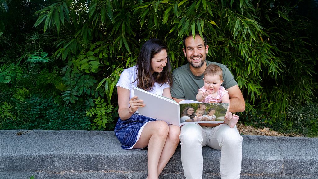 Eltern blättern mit einem Baby im Arm ein Fotobuch von Journi durch 
