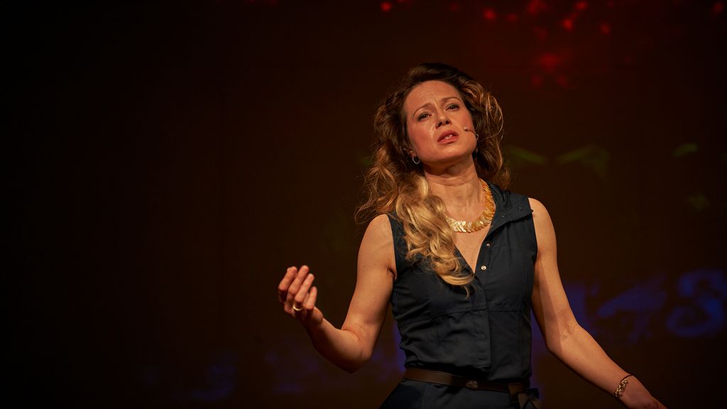 Eine Frau steht auf einer dunklen Bühne und gestikuliert ausdrucksstark im Licht