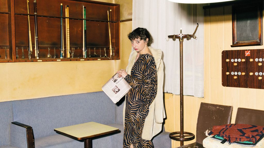 Frau steht mit einer Zeitung in der Hand auf der Bühne im Wohnzimmerstil