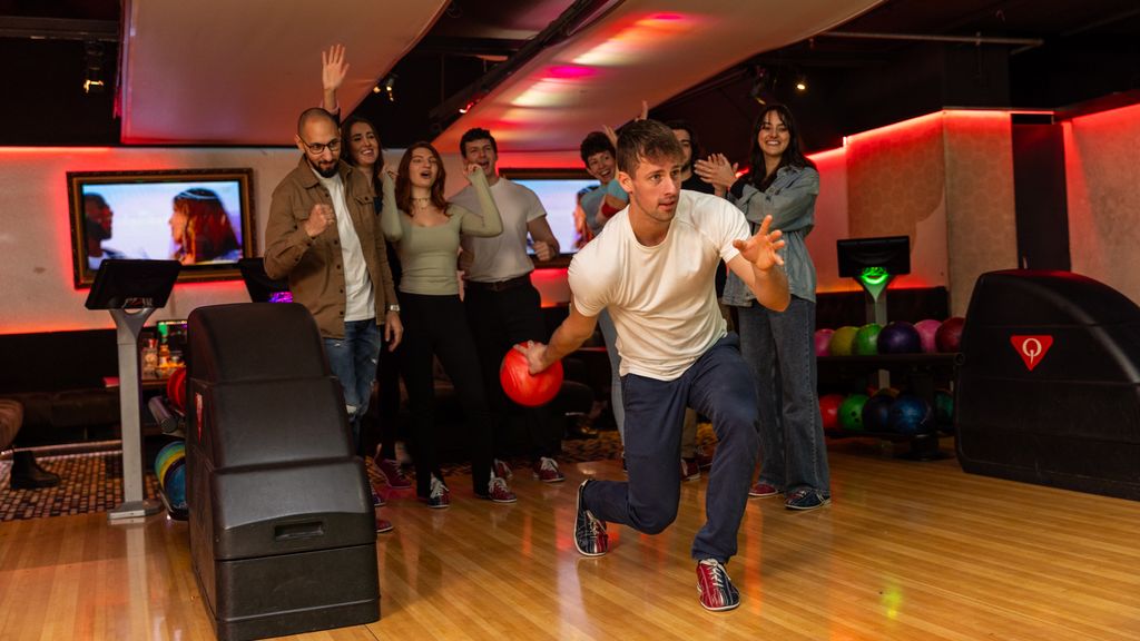 Ein Mann, der gerade die Bowlingkugel schwingt und im Hintergrund eine Gruppe, die ihn anfeuert. 