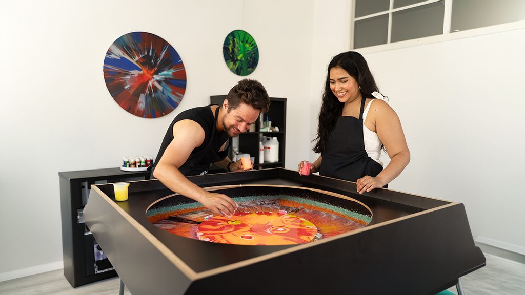 Zwei Personen gestalten gemeinsam ein buntes Kunstwerk an einer rotierenden Leinwand