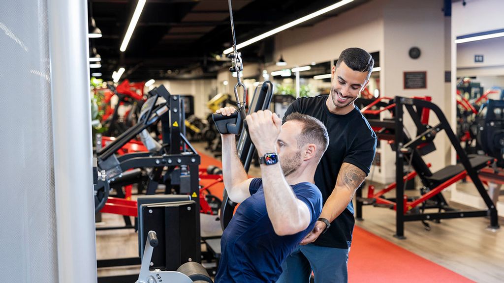 Personal Trainer unterstützt Mann bei einem Fitness Gerät im Fitness Studio