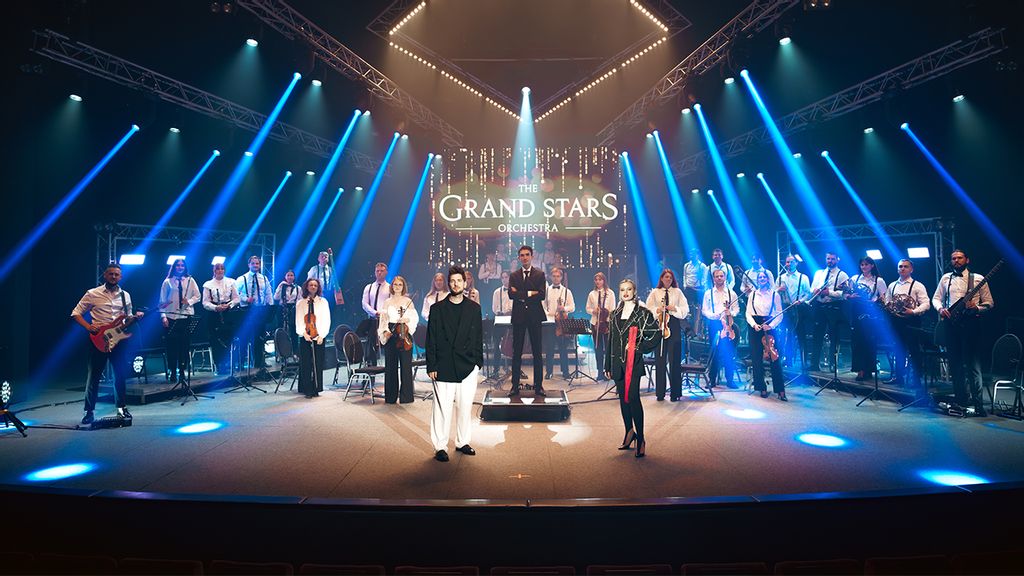 Zwei Sänger*innen stehen auf einer Bühen vor einem Dirigenten und einem Orchester