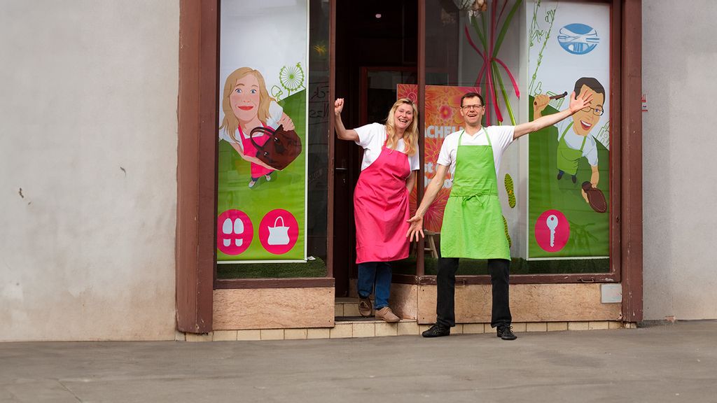 ein Mann in grüner Schürze und eine Frau in rosa Schürze vor dem Reparaturladen