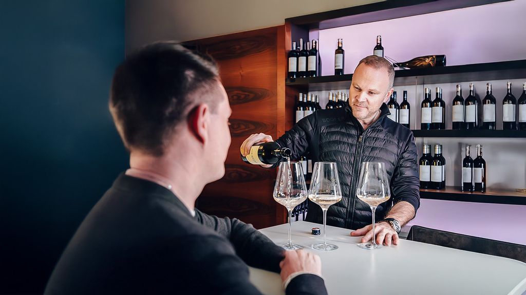 Ein Mann schenkt hinter der Theke Wein in Weingläser, während ein Gast dabei zuschaut.