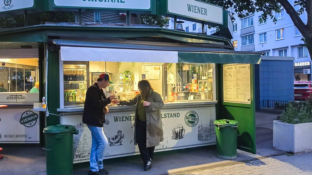 Zwei Personen essen an einem Würstelstand bei Dämmerung