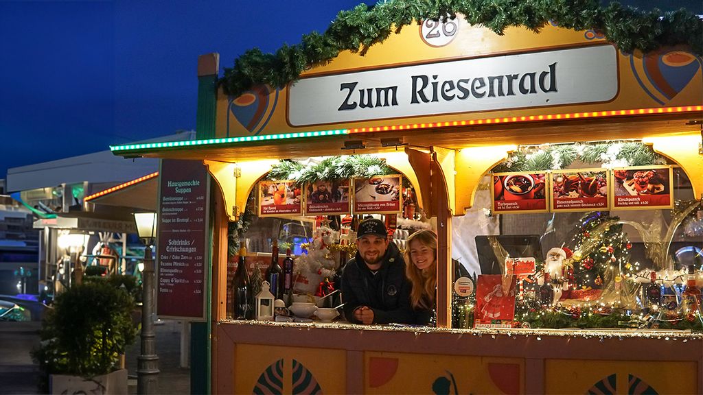 Weihnachtsmarkt-Stand bei Nacht mit Verkäufer*innen hinter dem Tresen.