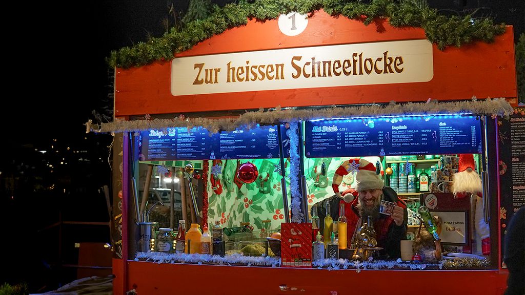 Weihnachtsmarkt-Stand „Zur heißen Schneeflocke“ bei Nacht mit Verkäufer hinter dem Tresen.