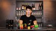 Barkeeper steht hinter einer Bar und präsentiert mehrere bunte Cocktails 
