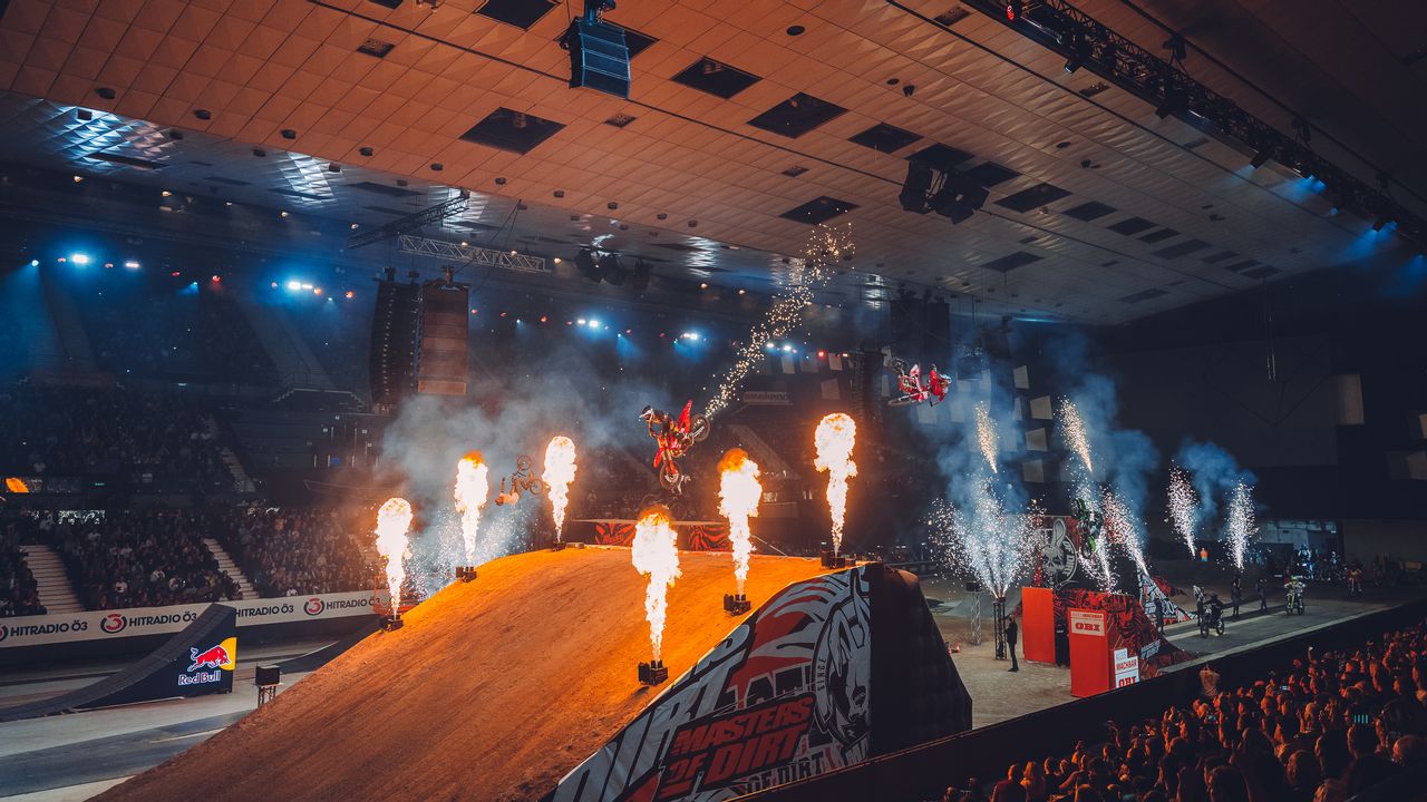 Motocross-Biker springt über eine Schanze und dreht sich in der Luft, rundherum sieht man Feuerwerk