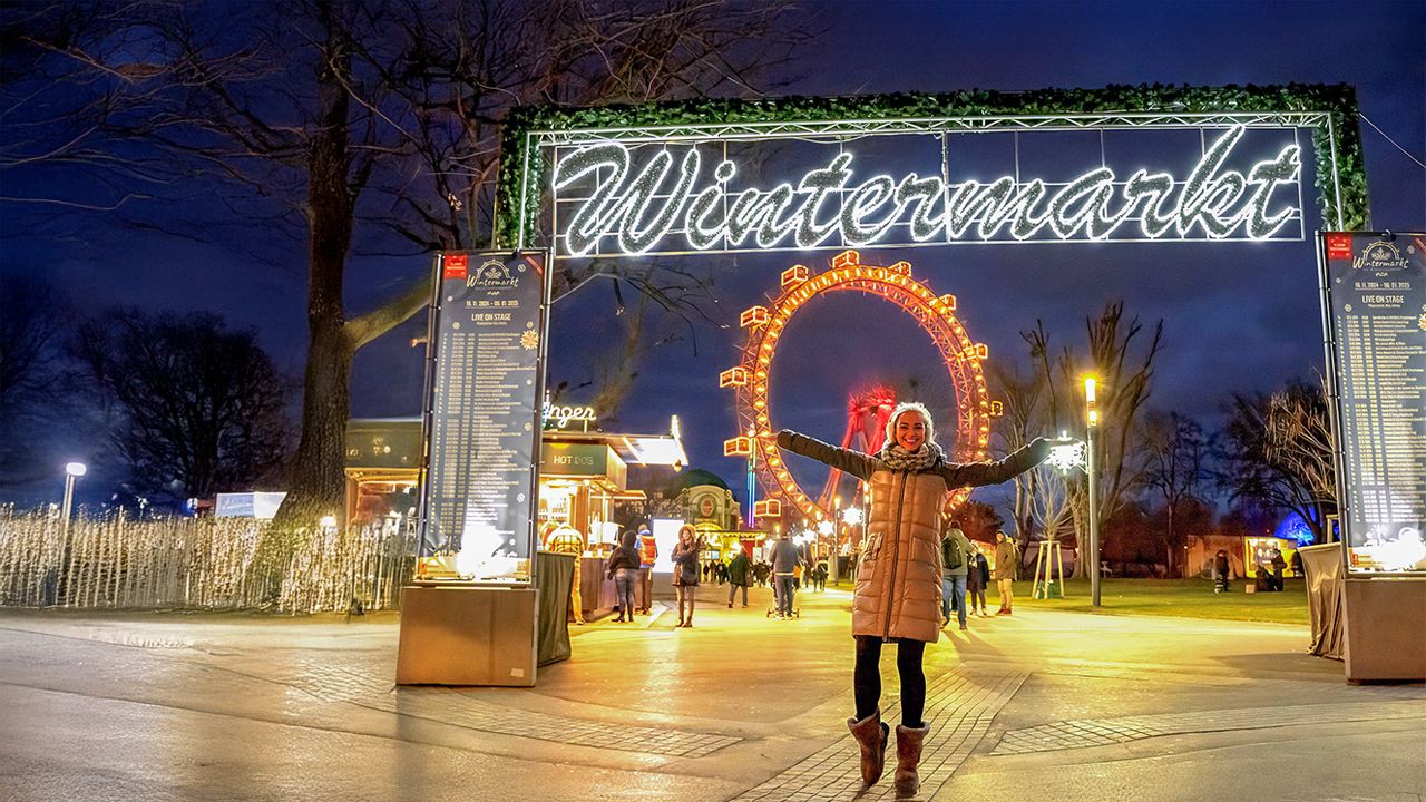 © Julius Silver wintermarkt.at Eine Frau steht vor dem beleuchteten Eingang vom Wintermarkt und im Hintergrund das Riesenrad