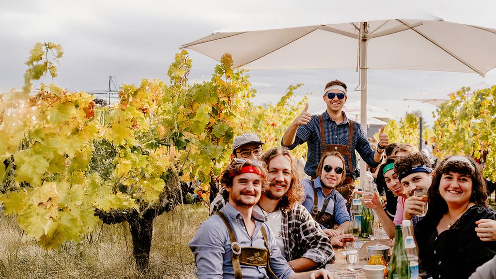 Eine Gruppe von Personen, die an einem Heurigentisch sitzen und trinken, im Hintergrund Weinreben.