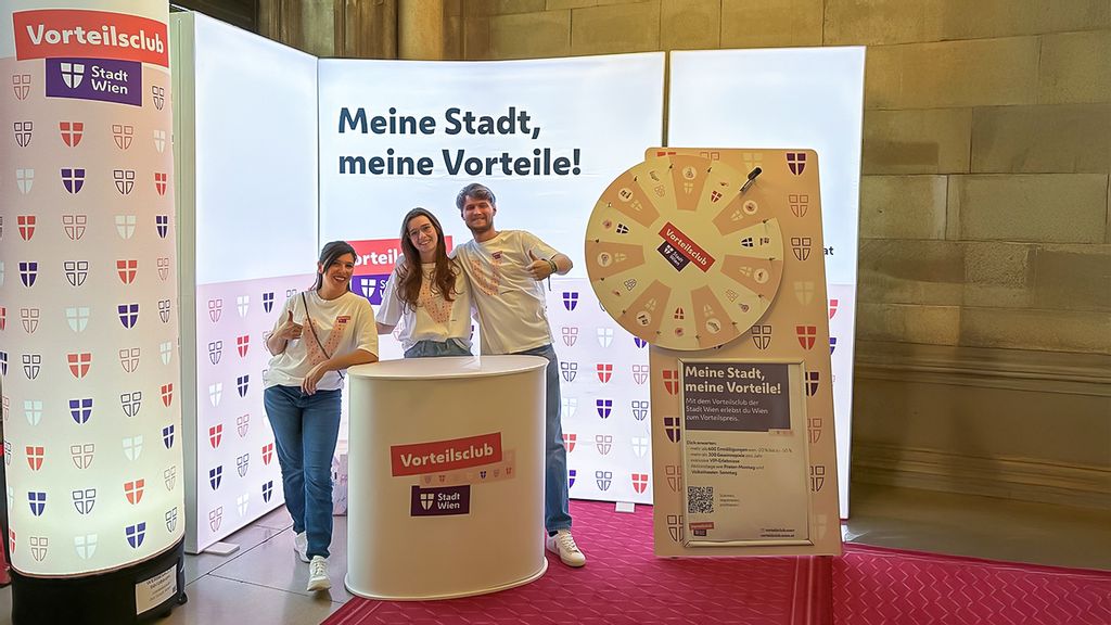 3 Menschen im Vorteilsclub T-Shirt vor dem Vorteilsclub Messe-Stand mit Glücksrad und Info-Desk 