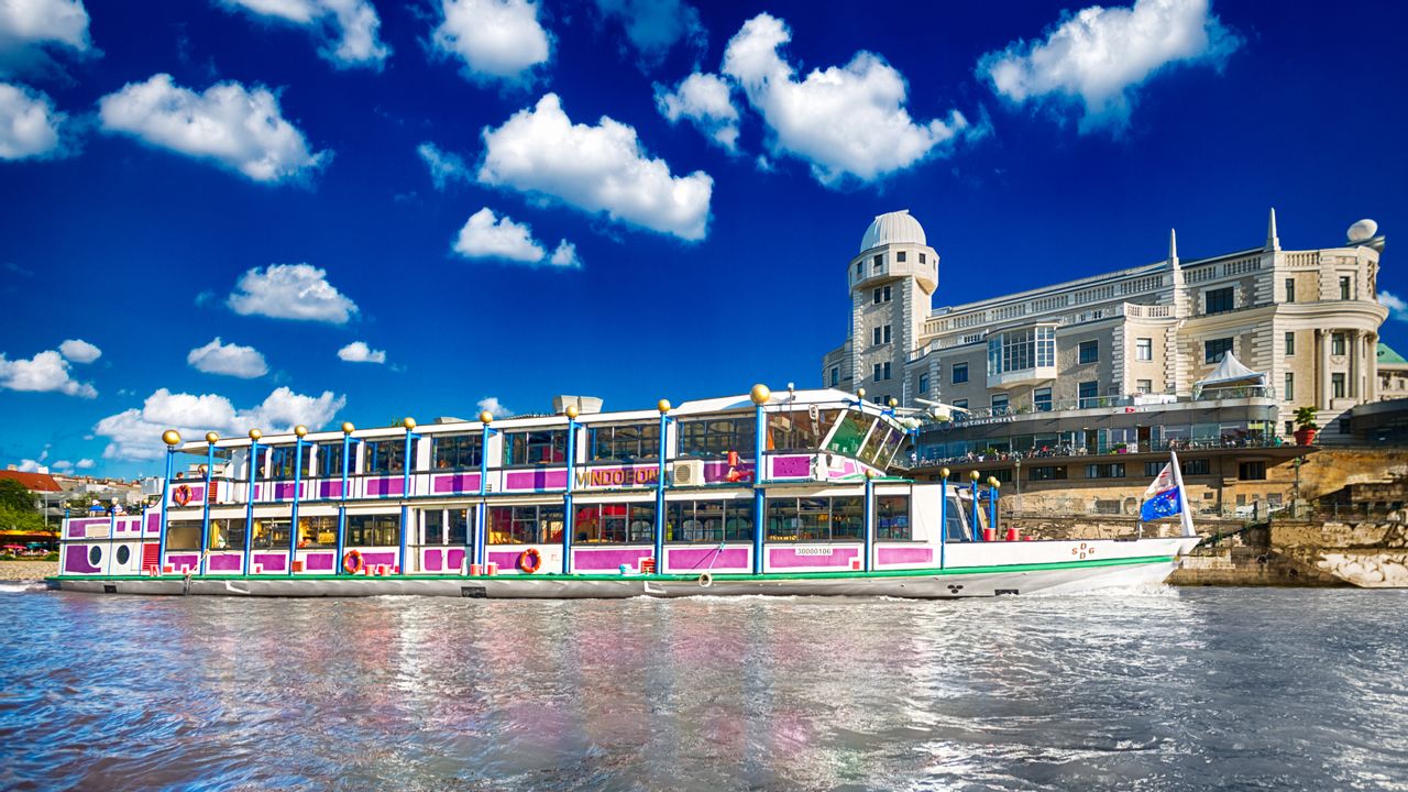 Ein buntes Schiff fährt den Donaukanal entlang, im Hintergrund sieht man die Urania 