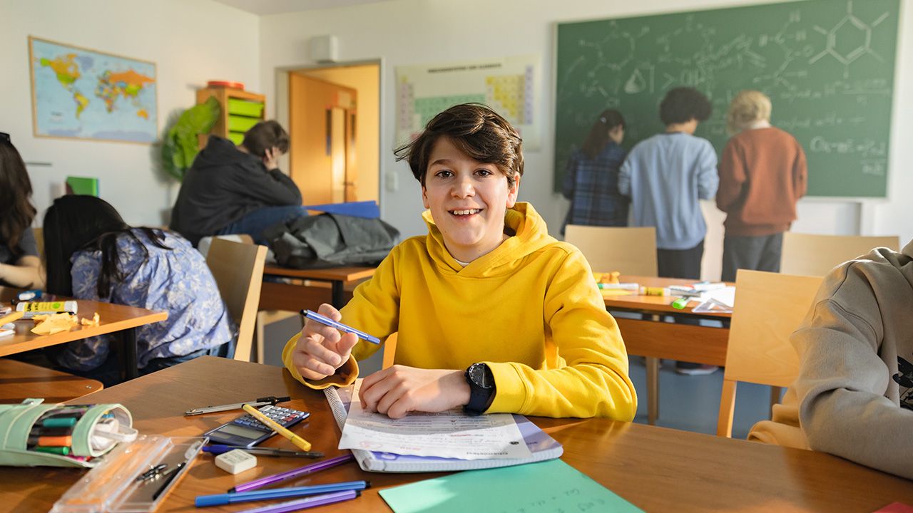 Ein Junge sitzt in einem Klassenzimmer auf einem Tisch, auf dem Papier und Stifte liegen