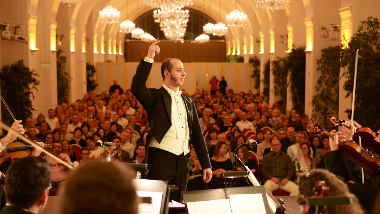 Ein Dirigent und Orchester auf einer Bühne vor Publikum in einem prunkvollen Konzertsaal