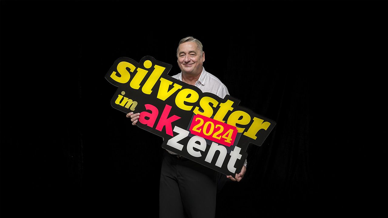 Der Kabarettist Andreas Vitasek hält den Schriftzug "Silvester im Akzent 2024" in der Hand