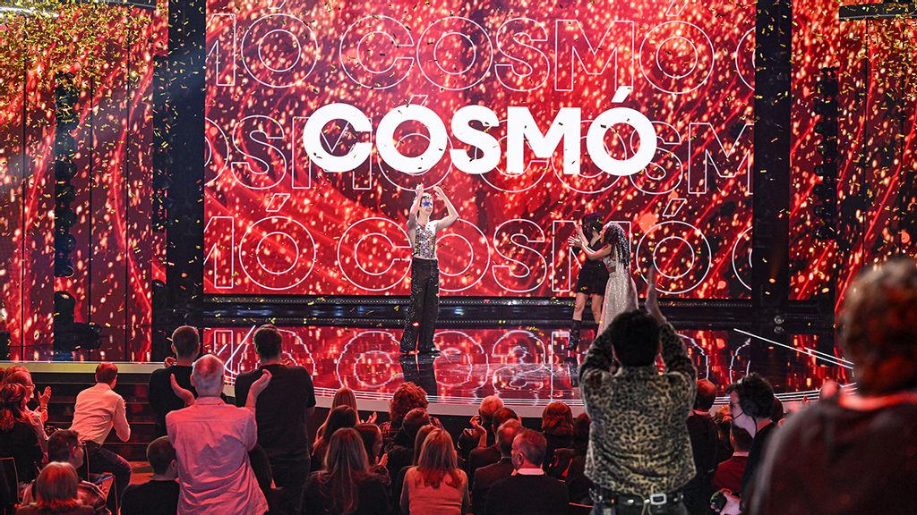 Ein Mann auf einer Bühne die rot beleuchtet ist und wo im Hintergrund COSMÓ steht