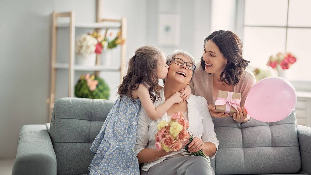 Ein Mädchen gibt ihrer Oma einen Kuss auf die Wange und die Mutter steht mit einem Geschenk daneben
