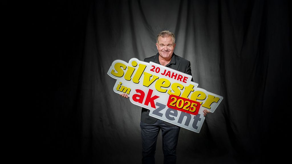 Cornelius Obonya hält ein Schild in der Hand worauf "20 Jahre Silvester im Akzent" steht