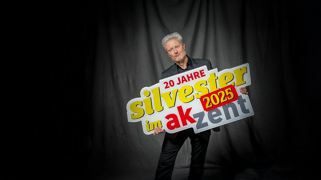 Florian Scheuba hält ein Schild in der Hand worauf "20 Jahre Silvester im Akzent" steht