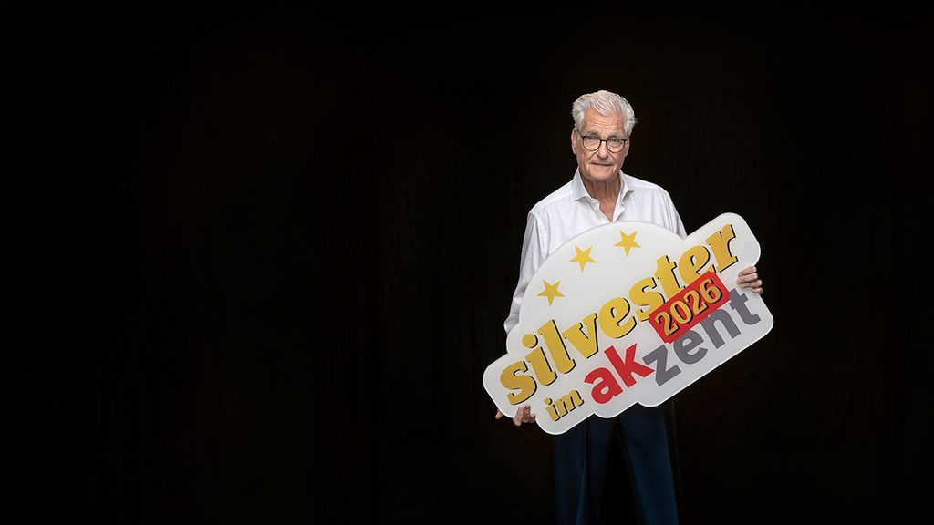 Schauspieler Sky du Mont hält ein Schild mit der Aufschrift: silvester im Akzent 2026, in den Händen