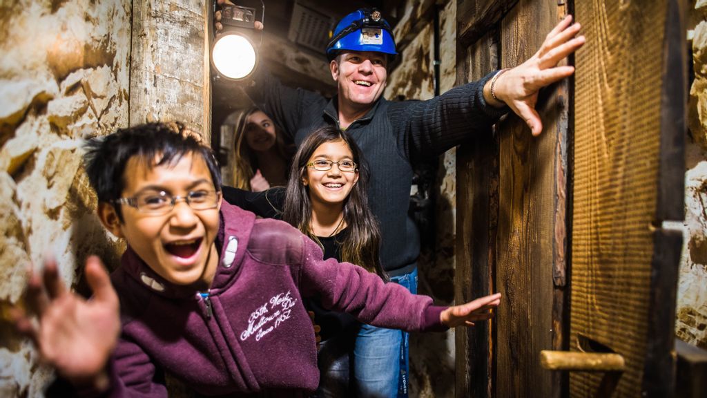 Zwei lachende Kinder und ein Mann mit Taschenlampe in der Hand und Helm am Kopf in einem Keller