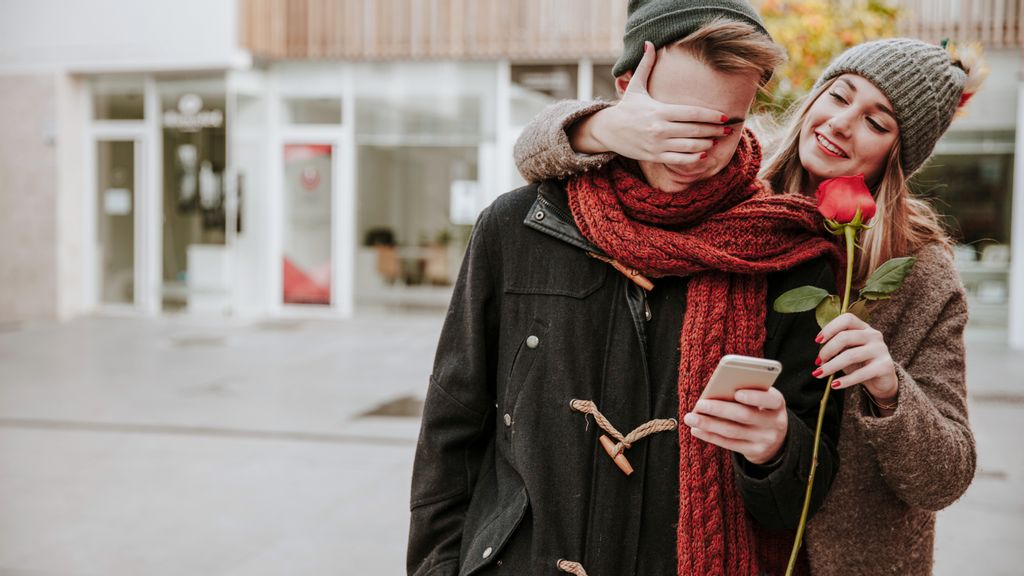 Eine Frau steht hinter einem Mann mit einer Rose in der Hand und hält ihm die Augen zu