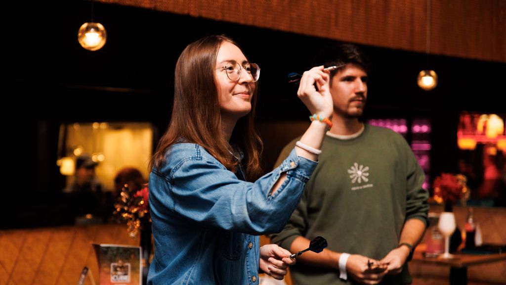 Eine Frau mit einem Dartpfeil in der Hand und im Hintergrund steht ein Mann und sieht zu