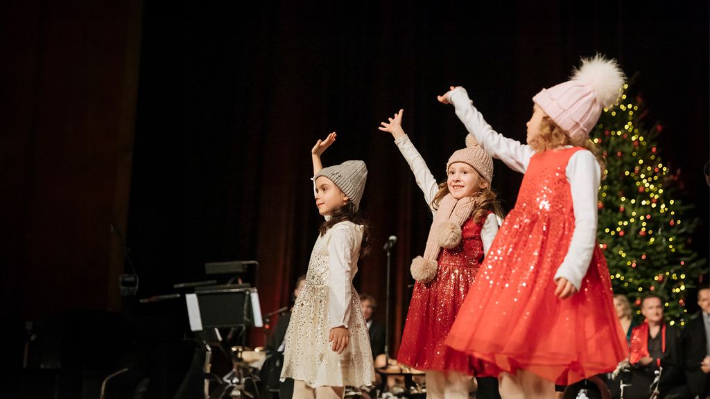 Drei winkende Kinder in weihnachtlichen Kleidern auf einer Bühne