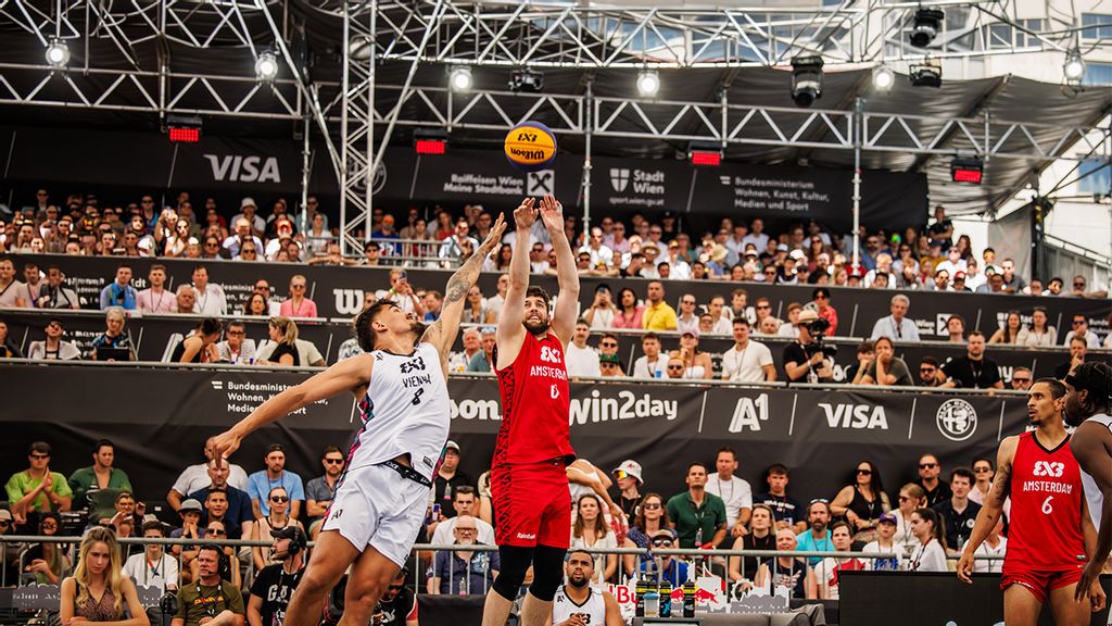 Basketballspieler mit einem Ball am Spielfeld dahinter Tribüne mit Zuseher*innen